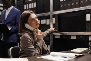 Inspector explaining police report using landline phone, working overtime at criminal investigation in arhive room. Private detective analyzing crime scene evidence searching for new clues photo