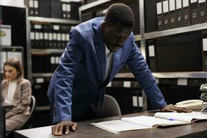 Bookkeeper working at bureaucracy report, analyzing administrative files late at night in storage room. African american businessman searching for bookkeeping record in corporate depository photo