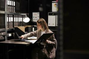 Investigator reading criminology files, working late at night at criminal case in arhive room. Police officer analyzing crime scene evidence, checking documents with confidential victim information photo