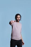 Cool arab man showing neutral thumb sign with hand and looking at camera. Young person raising eyebrow and making satisfaction normal level gesture with finger on blue background photo