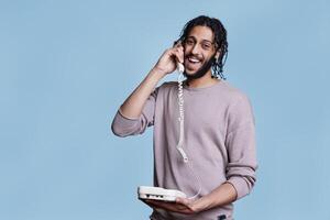 Smiling arab man talking on landline phone and looking at camera. Cheerful person answering retro telephone call, having fun conversation and laughing with carefree facial expression portrait photo