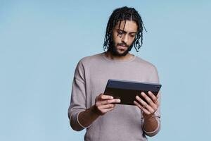 Concentrated arab man holding digital tablet and reading social media online. Focused young person surfing internet and browsing pages while using portable electronic gadget photo