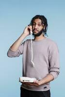 Arab man having conversation on landline phone and looking at camera. Confused person answering call and listening to collocutor while talking on retro telephone portrait photo