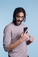 Excited arab man checking message on smartphone and showing winner gesture with clenched fist portrait. Person using mobile phone and looking at camera with happy expression photo
