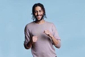 Arab man smiling and moving hands with clenched fists, dancing while listening to music portrait. Young person in good mood having fun and looking at camera with cheerful facial expression photo