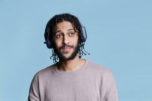 Arab man enjoying music wearing wireless headphones and looking away with dreamy facial expression. Pleasant young person listening to song in earphones on blue background photo