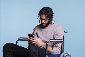 Arab man sitting in wheelchair while scrolling social media on smartphone. Young person with physical impairment sending online message and browsing internet on mobile phone photo