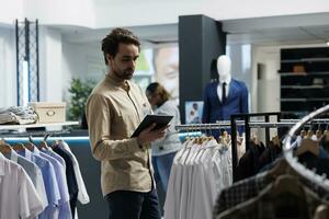 Department mall employee managing inventory with digital tablet, examining and organizing apparel. Clothing store young man worker checking merchandise in stock while working photo