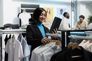 Clothing store smiling manager using digital tablet to check stock availability while browsing through apparel rack. Shopping center boutique asian woman employee managing inventory photo