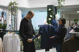 Shopping center boutique woman worker holding jacket on hanger, assisting arab customer in choosing outfit. Clothing store asian employee consulting client, helping with purchase photo