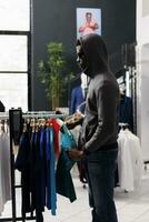 African american man looking aside while robbing clothing store, checking racks with trendy clothes. Thief trying to steal fashionable merchandise, wearing sunglasses and hood not to be recognized photo