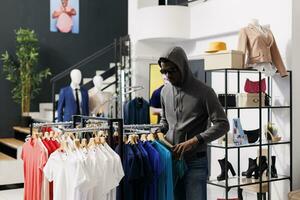 africano americano ladrón robando casual camiseta, mirando alrededor a ver Si alguien acecho él en moderno boutique. hombre robando ropa almacenar, vistiendo capucha y Gafas de sol. crimen concepto foto