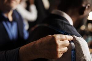 Close up of professional experienced suitmaker doing fitting length estimations on sartorial piece using measurement tape. Meticulous couturier helping client with custom handmade elegant suit photo