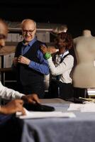 Cheerful meticulous seamstress doing precise additional needlework needed on sartorial piece worn by elderly client. Skilled african american tailor helping customer with bespoke suit comission photo