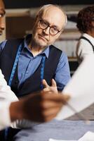 Close up of master tailor checking aspiring fashion design apprentice sartorial garment sketches in atelier workspace. Couturiers working on bespoken client customization comission photo