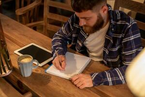 Young bearded businessman sits in cafe, home at table and writes in notebook, near lies tablet computer with black screen. Man is working, studying. photo