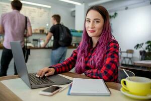 Young woman with pink hair with laptop computer sitting in cafe, intelligent female student working on net-book. photo