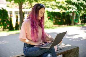 joven niña es estudiando en el primavera parque, sentado en el de madera banco y hojeada en su ordenador portátil foto