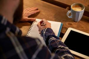Young bearded businessman sits in cafe, home at table and writes in notebook, near lies tablet computer with black screen. Man is working, studying. photo