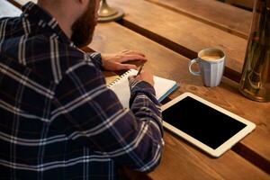 Young bearded businessman sits in cafe, home at table and writes in notebook, near lies tablet computer with black screen. Man is working, studying. photo
