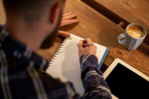 Young bearded businessman sits in cafe, home at table and writes in notebook, near lies tablet computer with black screen. Man is working, studying. photo