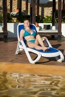 Young woman relaxing on the deck chair by the swimming pool at the resort photo