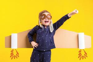 Freedom, girl playing to be airplane pilot, funny little girl with aviator cap and glasses, carries wings made of brown cardboard as an airplane photo