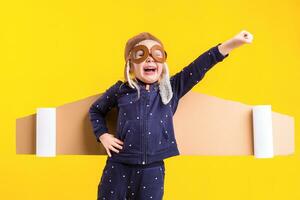 Freedom, girl playing to be airplane pilot, funny little girl with aviator cap and glasses, carries wings made of brown cardboard as an airplane photo