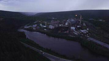 Top view of industrial city by river on summer evening. Clip. Beautiful landscape of industrial town by river among forest hills in evening. Highway passing by river with industrial plant and town photo