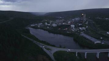 Top view of industrial city by river on summer evening. Clip. Beautiful landscape of industrial town by river among forest hills in evening. Highway passing by river with industrial plant and town photo