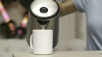 Close-up of young woman pouring hot water from electric kettle in kitchen. Stock footage. Modern kitchen design photo