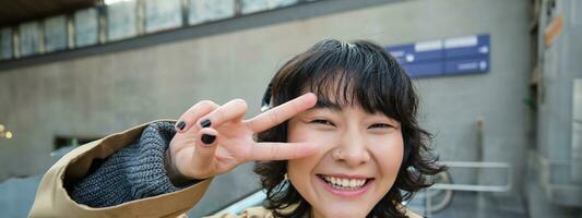 Positive korean girl in headphones, drinks coffee to go, shows peace, v-sign, stands on street and smiles happily photo