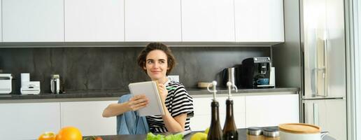 Portrait of young woman looking at cooking ingredients on kitchen counter and making notes, writing down recipes, thinking of meal for dinner, preparing vegetarian food photo