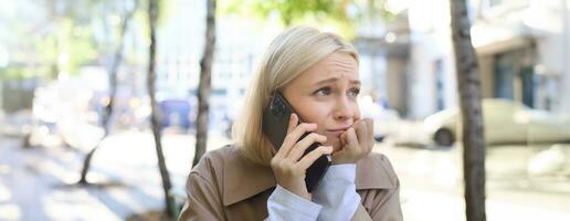 Image of young stressed, upset woman, talking on mobile phone with worried face expression, has difficult conversation over the telephone, sitting in outdoor cafe photo