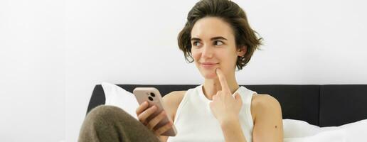 Close up portrait of smiling woman in bed, thinking, holding smartphone, looking aside with pleased smirking face, making decision, online shopping on mobile phone while spending time at home photo
