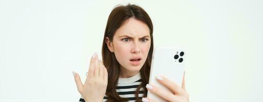 retrato de confundido, enojado mujer ceñudo, leyendo texto en teléfono inteligente, acecho vídeo en móvil teléfono con conmocionado, perplejo cara expresión, aislado en blanco antecedentes foto