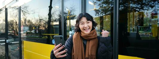 Enthusiastic asian woman, standing on bus stop with smartphone, looking at phone screen with amazed, triumphing face, winning, hear great news on video chat photo