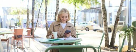 Image of young woman drinking coffee, sitting in outdoor cafe with smartphone, sending message, reading notification on mobile phone and smiling photo