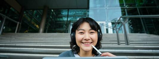 Close up of smiling asian girl feels inspired while draws on digital tablet, makes graphic design project, pencils user interface assignment, sits on stairs near campus photo