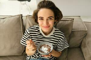 Close up of cute young female model, eating cereals with milk, enjoys her breakfast on sofa in living room, smiling and looking happy photo