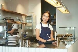 retrato de hermosa asiático niña sonriente, barista en café trabajando detrás encimera, utilizando tableta como pos Terminal, Procesando orden foto