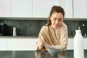 Portrait of beautiful woman at home, eating her breakfast, holding spoon, leaning on worktop and having cereals with milk photo