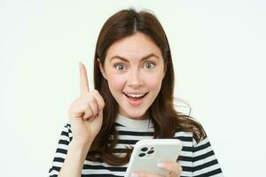 Got an idea. Smiling woman, raises her finger and holds smartphone, suggests something, makes eureka gesture, has a solution, white background photo