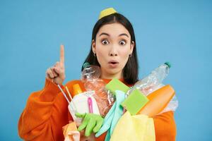 Happy young woman pointing up, holding plastic empty bottles and rubbish for recycling, showing eco banner, sorting station advertisement, blue background photo