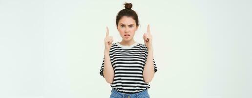 Portrait of shocked woman, raise eyebrows and points fingers up, looks complicated, puzzled by something, standing over white background photo