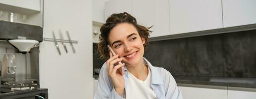 Portrait of young woman, 25 years old, holding smartphone, talking on mobile phone, sitting in kitchen at home photo