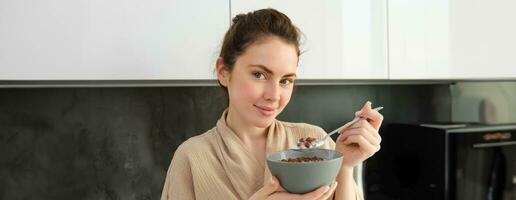 Good-looking brunette woman with bowl of cereals, eating meal for breakfast, wearing bathrobe, looking at camera and smiling photo