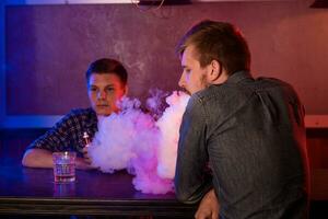 dos joven hombres fumar electrónico cigarrillos en un barra de vapor foto