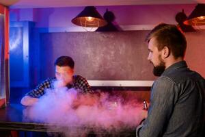 dos joven hombres fumar electrónico cigarrillos en un barra de vapor foto