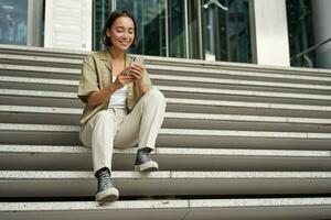 Cellular technology and people. Young happy asian girl sits with smartphone in front of building. Woman using mobile phone, smiling while looking at screen photo